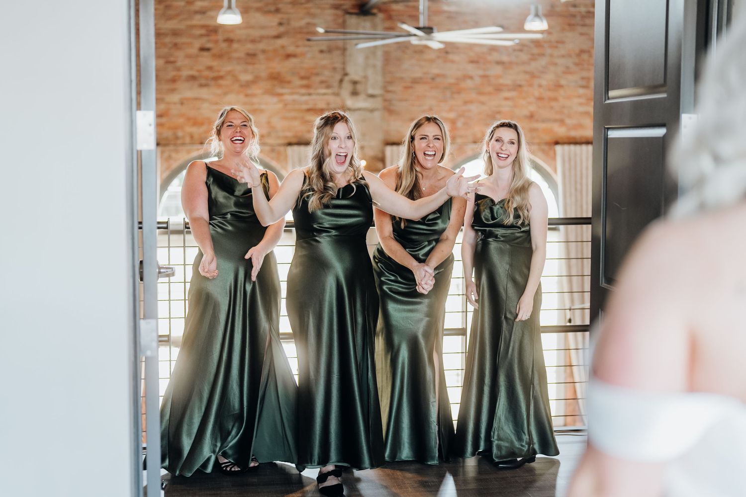 Bridesmaids wearing elegant sage green dresses pose together in a rustic brick-walled venue.