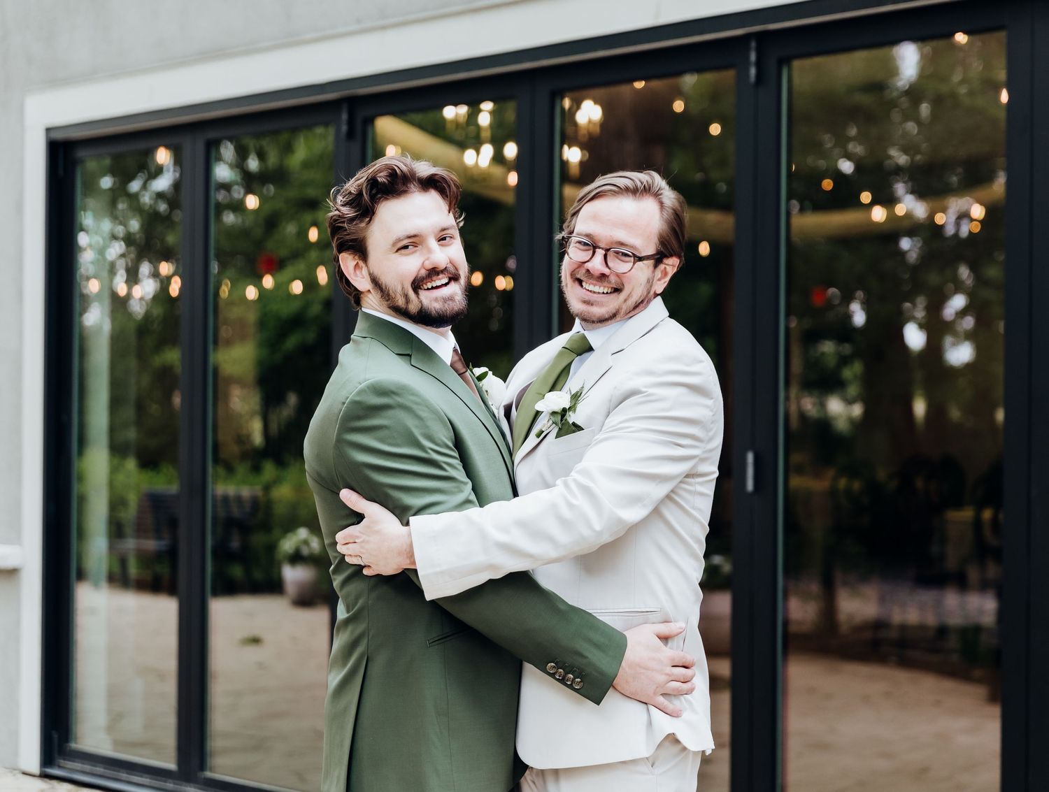 Wedding party members in white suits share laughter and drinks outside a venue with glass windows.