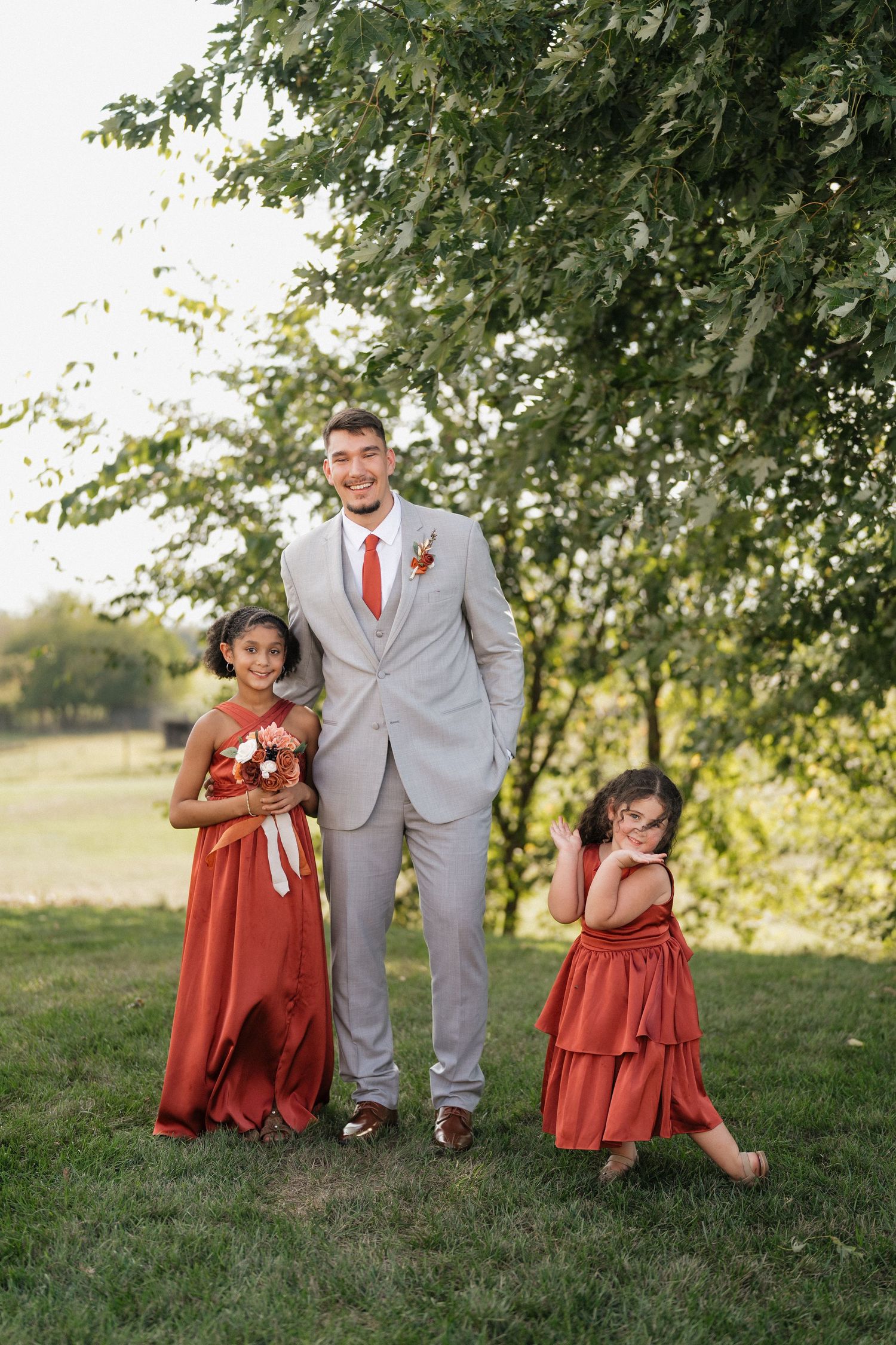 Wedding party members in grey suits and rust-colored dresses pose together under trees in an outdoor setting.
