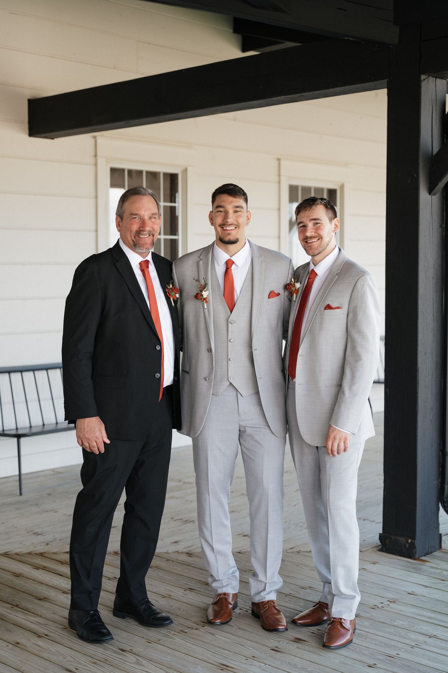 Three men in formal attire with red accents pose together on a covered porch.
