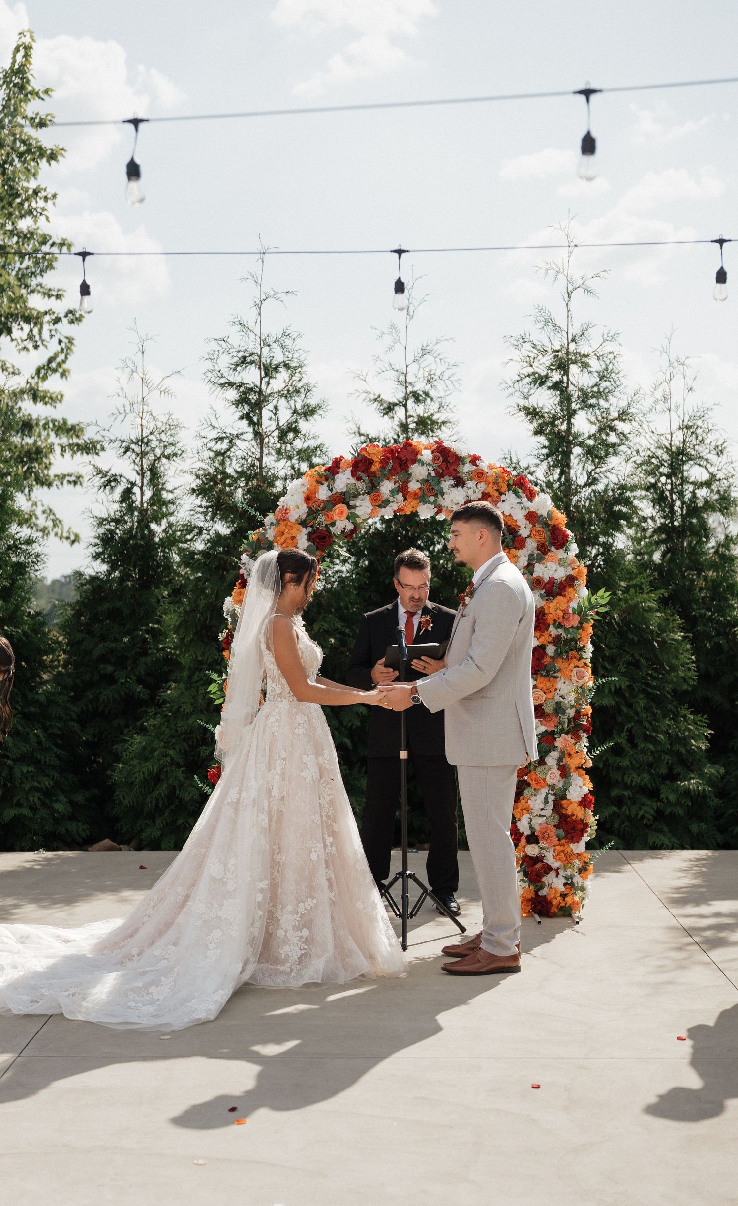 Wedding ceremony under floral arch with coral and white flowers against cloudy sky.