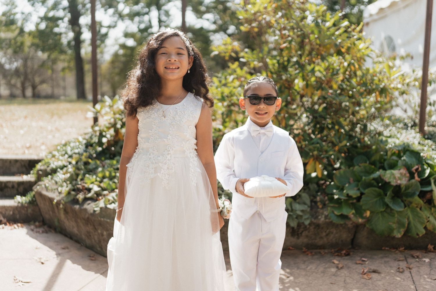 A group of children in white dresses stand in a garden setting during a formal outdoor event.