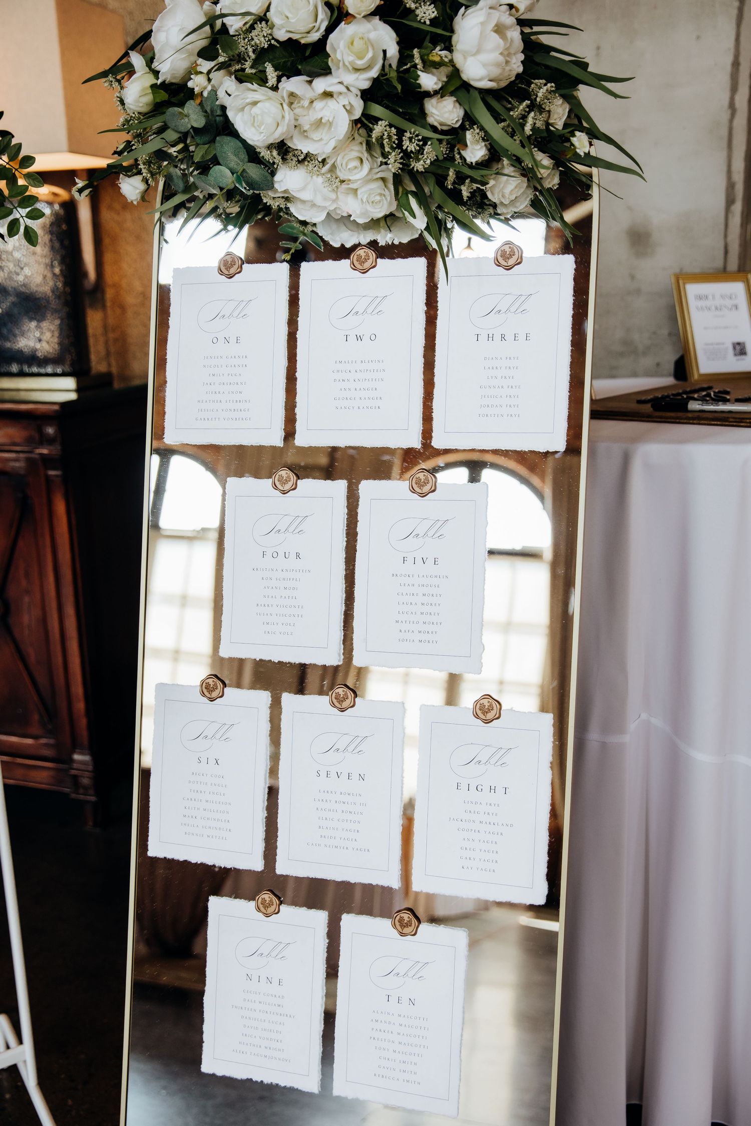 Wedding seating chart display with white cards and floral decorations hanging against white curtains.