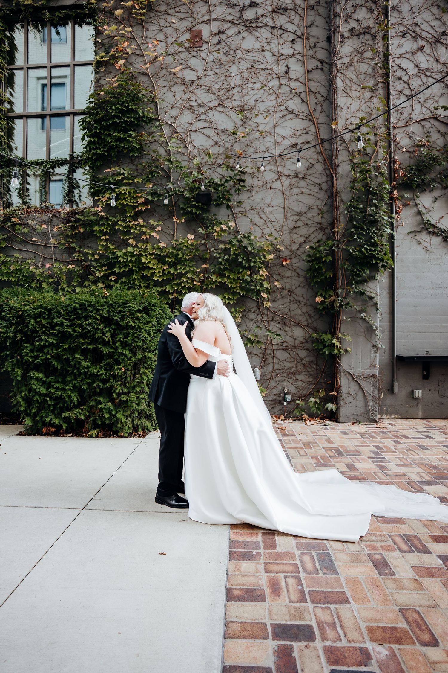 Bride in flowing white gown and groom share intimate moments against ivy-covered stone wall.