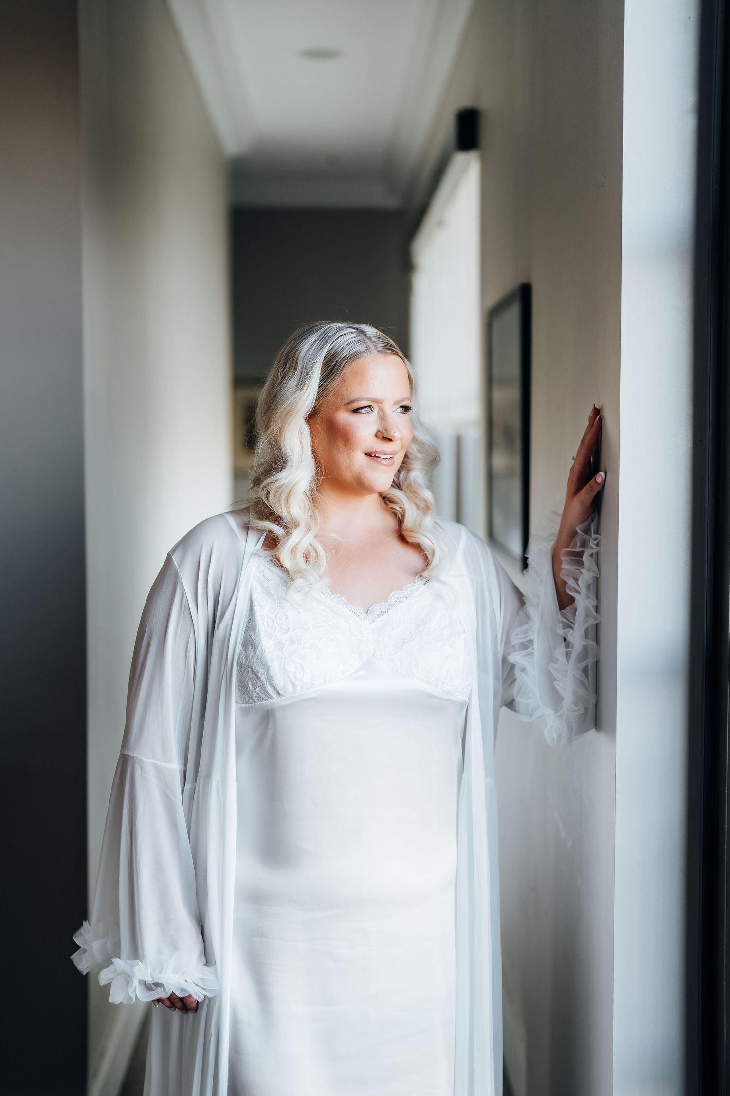 Bride in white silk robe stands by window during wedding morning preparation.