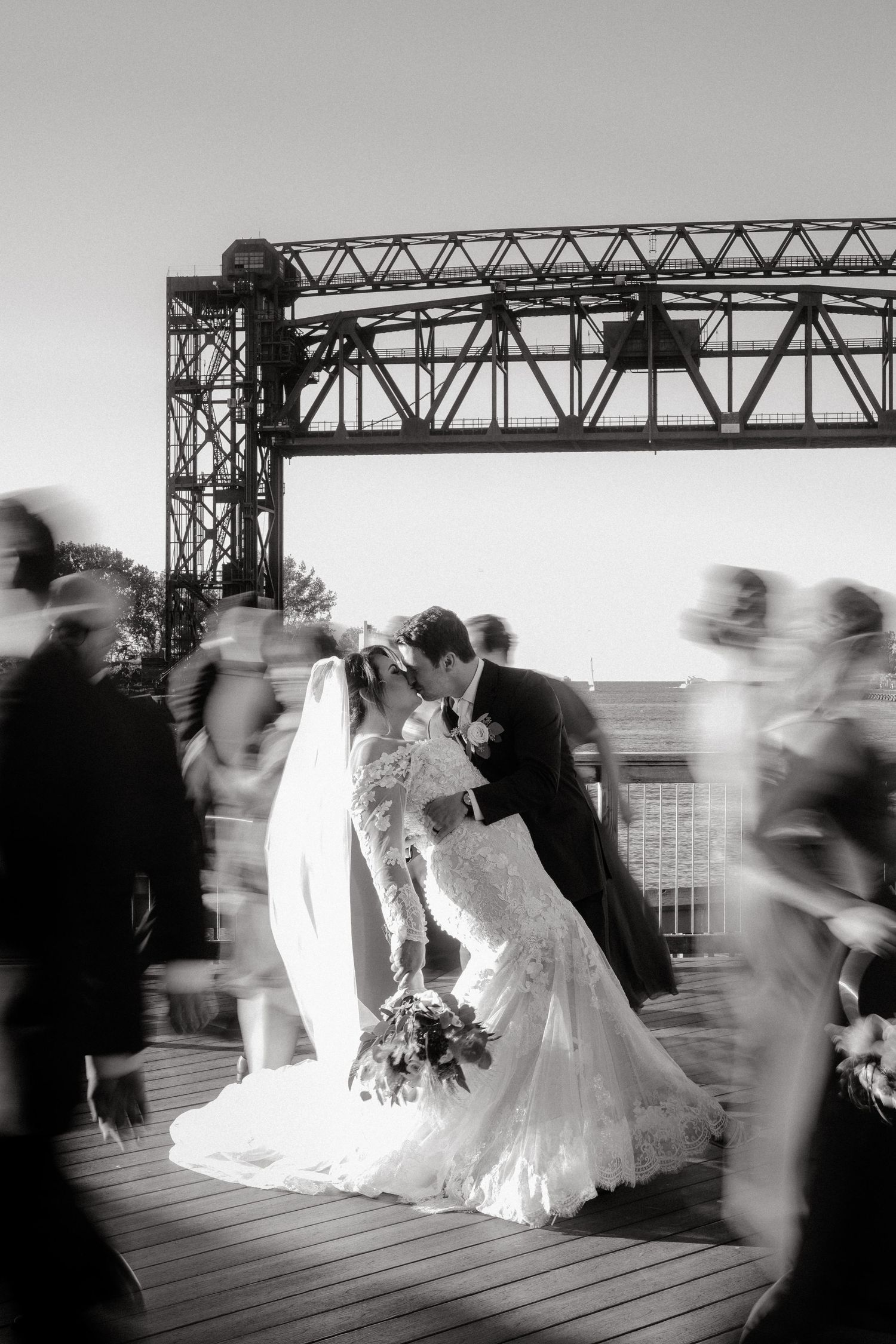 Artistic black and white wedding photo under an industrial bridge with motion-blurred guests.