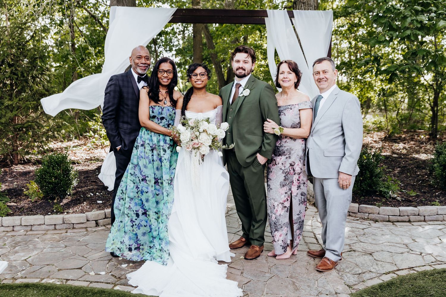 Wedding party poses together outdoors on stone patio surrounded by greenery and trees.