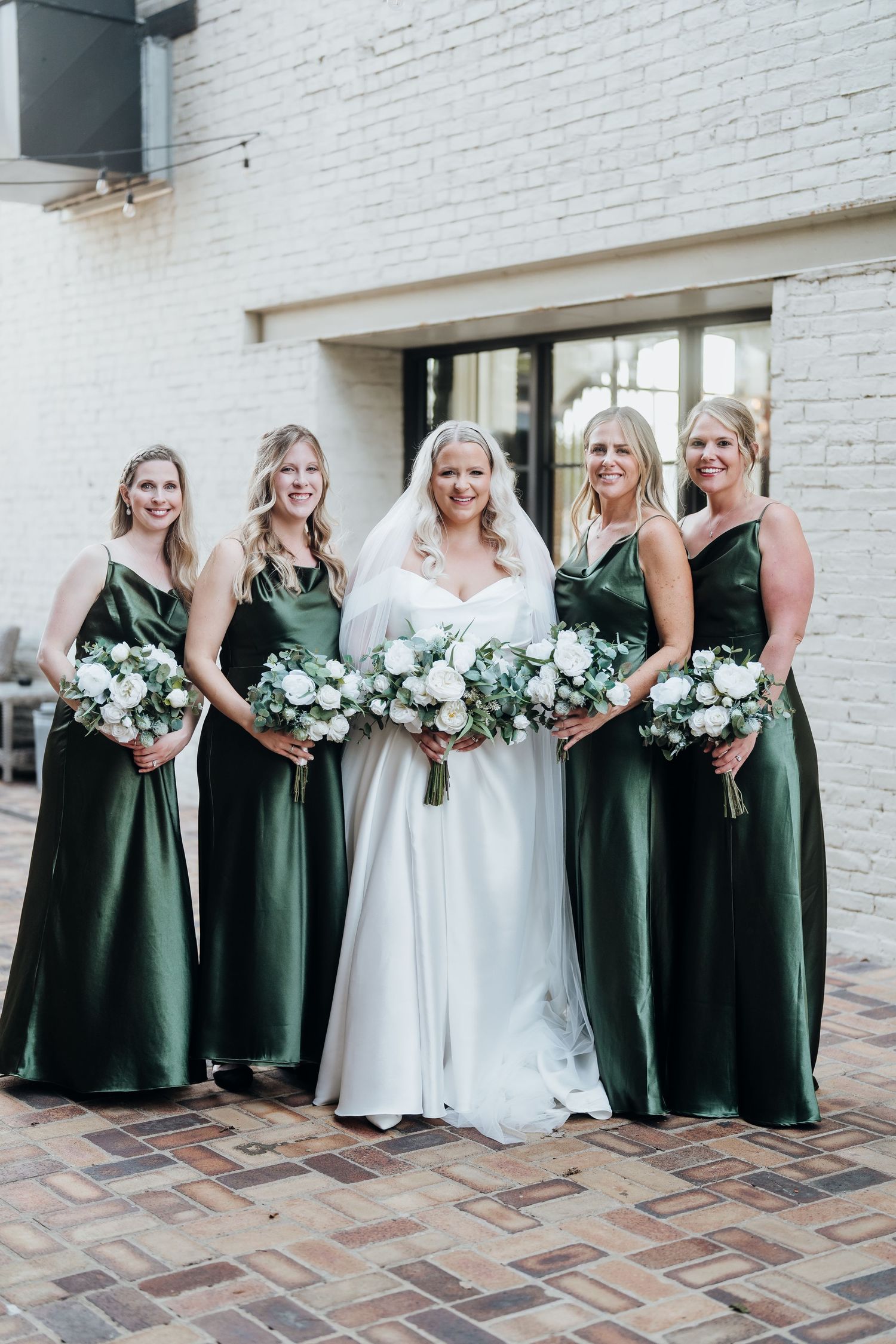 Wedding party poses playfully with bridesmaids in dark green dresses against white brick backdrop.