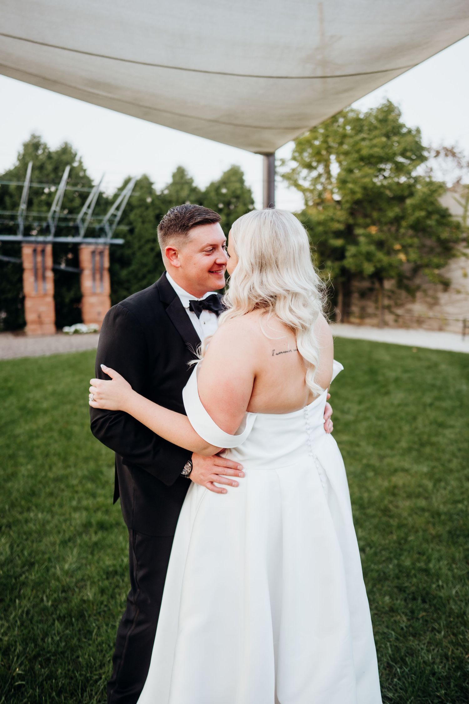 A couple in wedding attire embraces on a grassy lawn with brick buildings in the background.