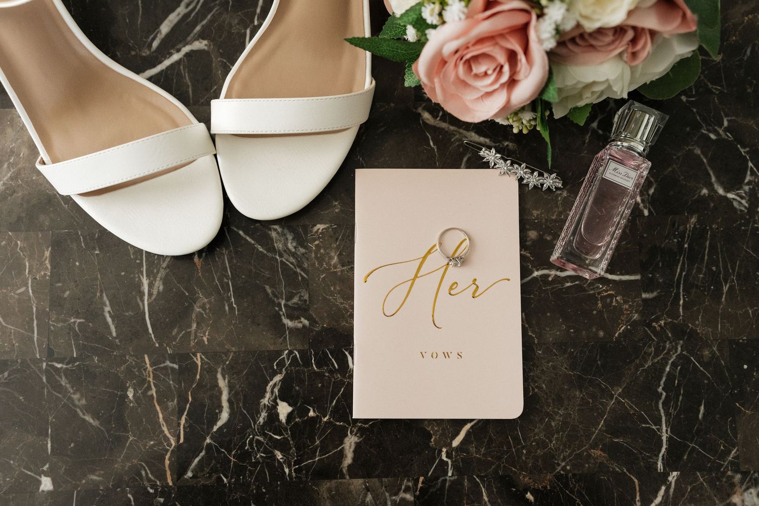 Elegant wedding details including pink roses, invitations, rings, and perfume bottle arranged on dark marble surface.