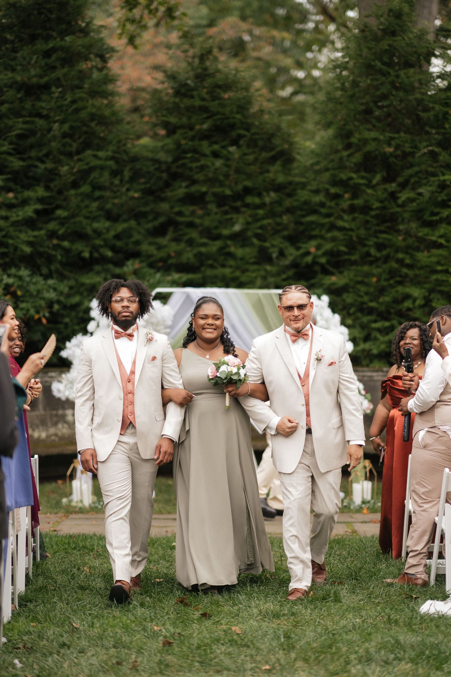 A wedding party in white and beige attire poses joyfully together on a grassy lawn surrounded by evergreen trees.