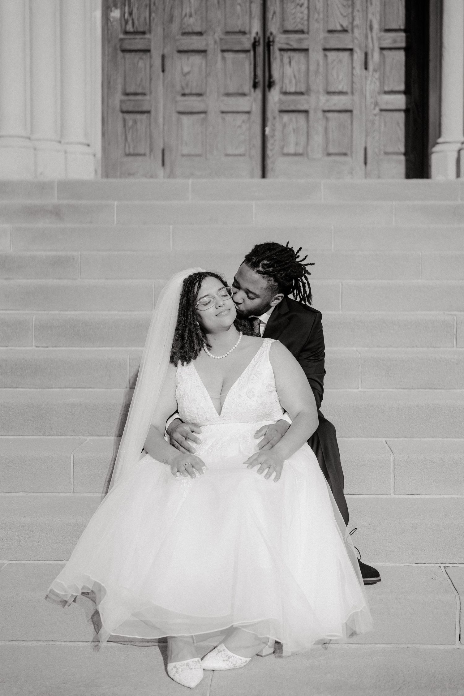A couple poses on church steps in elegant wedding attire for a black and white portrait.
