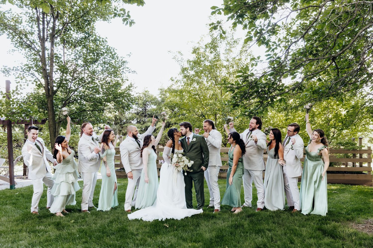 Wedding party in white and mint green attire poses together on grass in front of wooden fence.