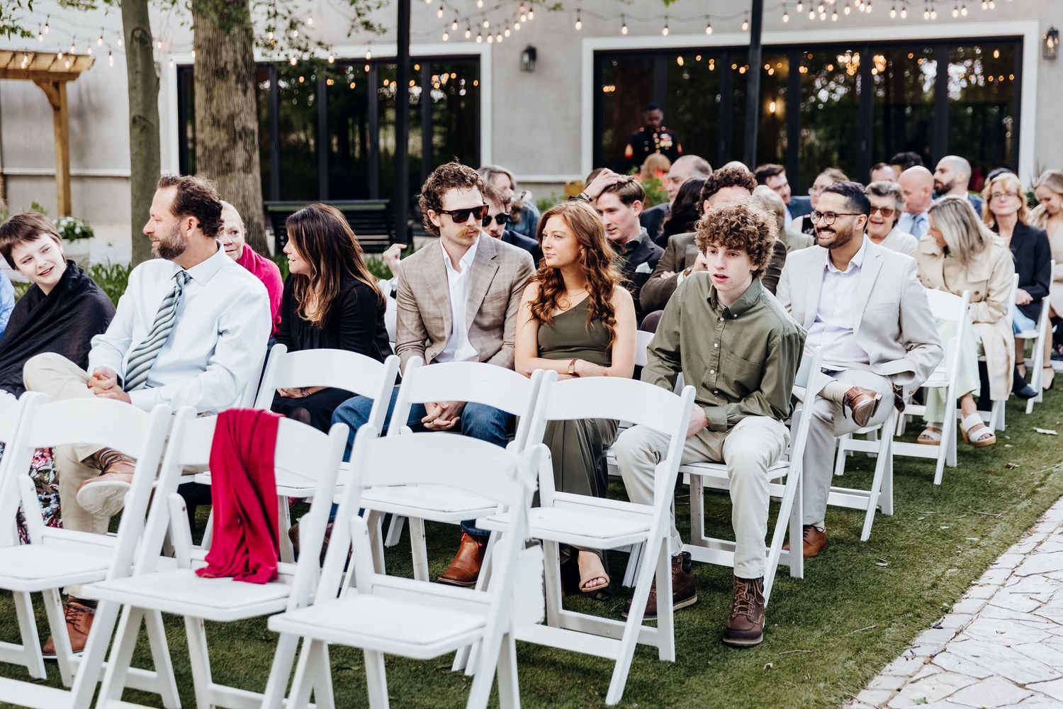 Outdoor wedding ceremony setup with rows of white chairs and guests seated under trees.