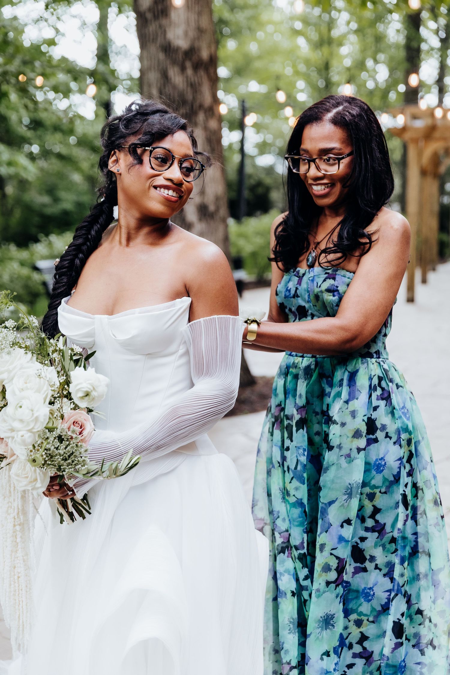 A bride in a white wedding dress stands with a bridesmaid wearing a blue floral gown in an outdoor garden setting.