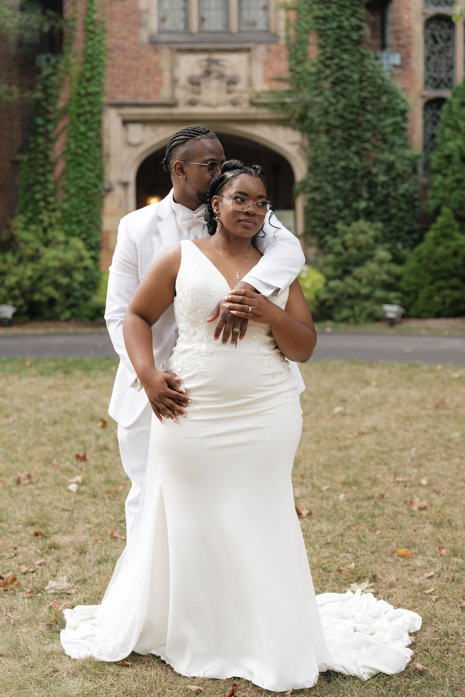 A joyful wedding couple embraces on a grassy lawn in front of a brick building with ivy-covered walls.