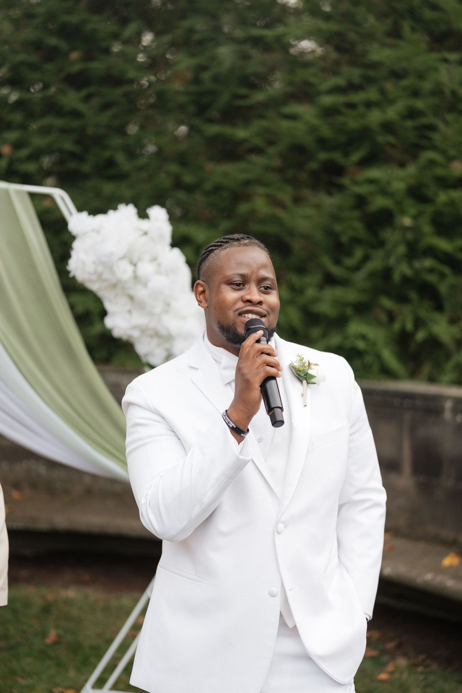 A person in a white suit and tie stands against a green backdrop at an outdoor event.