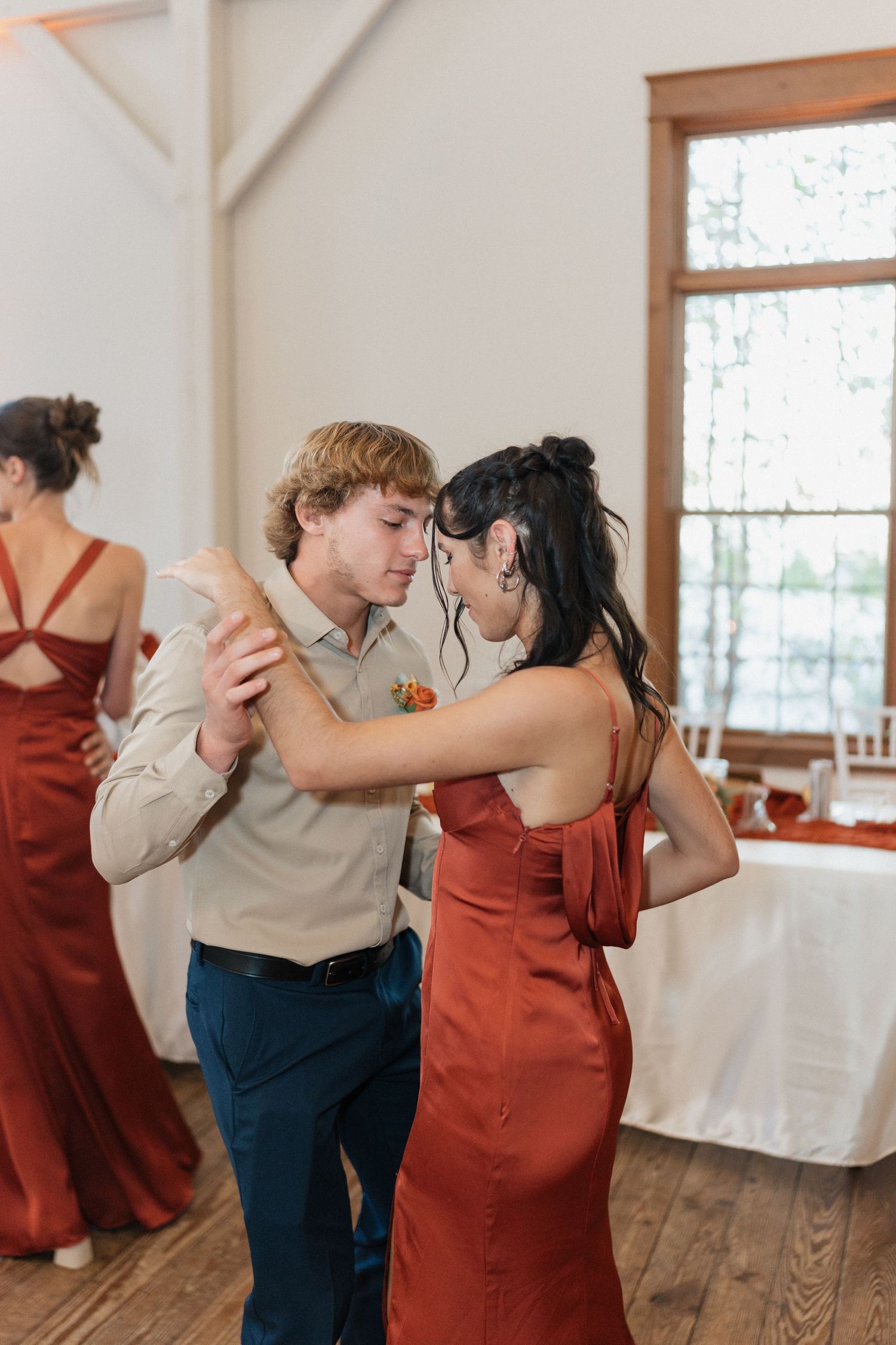 Wedding party dancing in indoor venue with wooden floors and white walls.