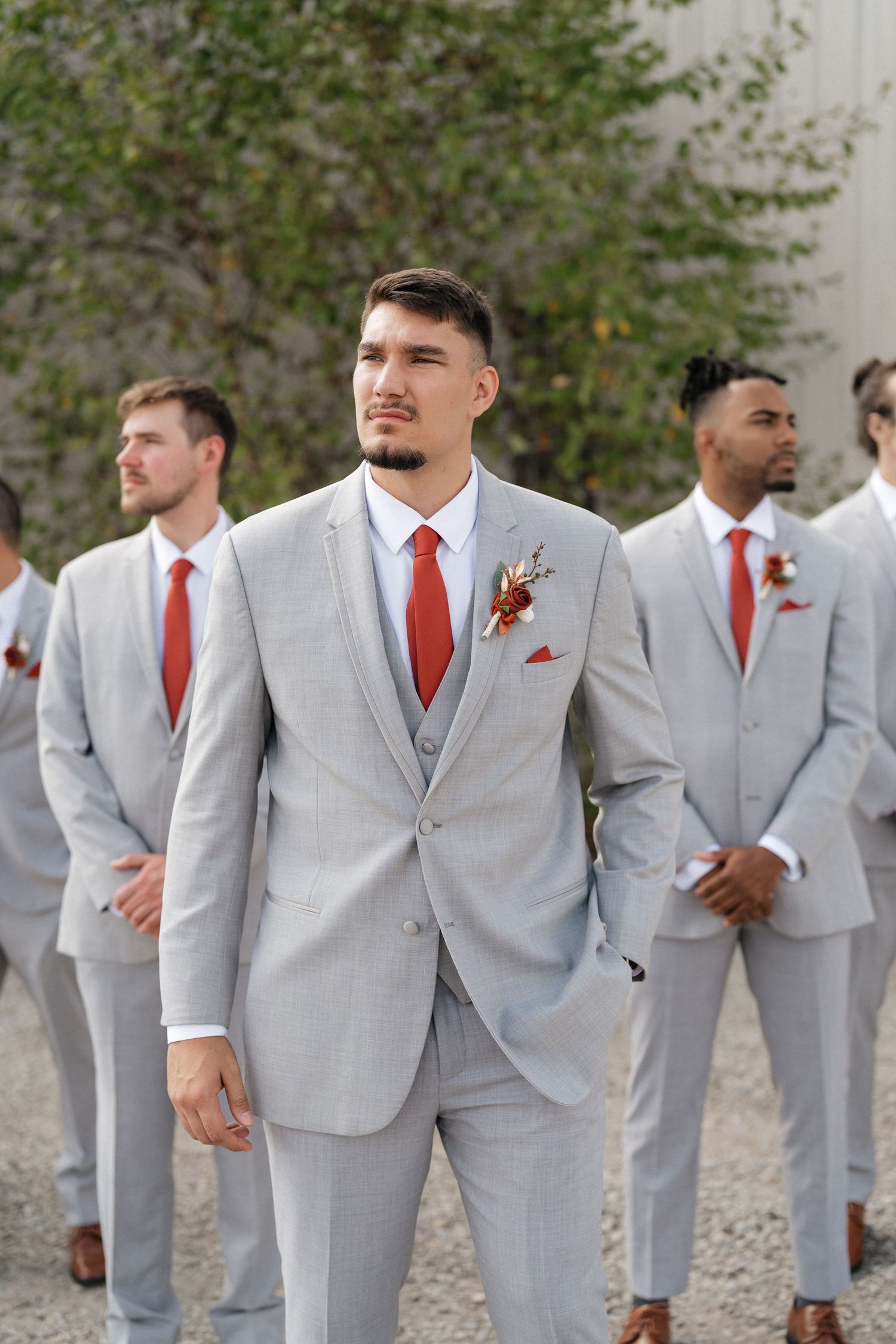Groomsmen in matching light gray suits and red ties stand together in a line outdoors.