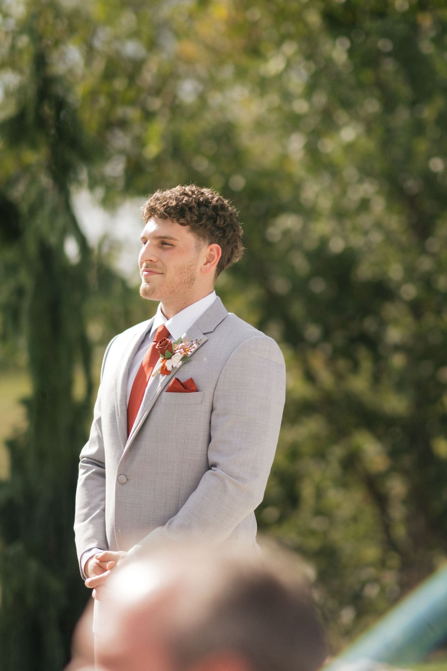 Groomsman in light gray suit with coral tie watching ceremony outdoors.