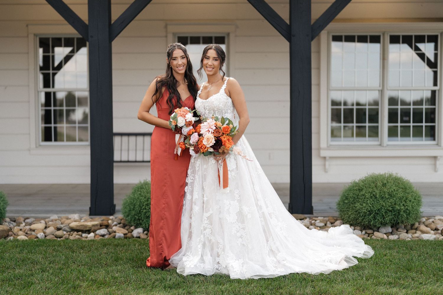 Two people pose outdoors in wedding attire holding vibrant orange and pink bouquets.