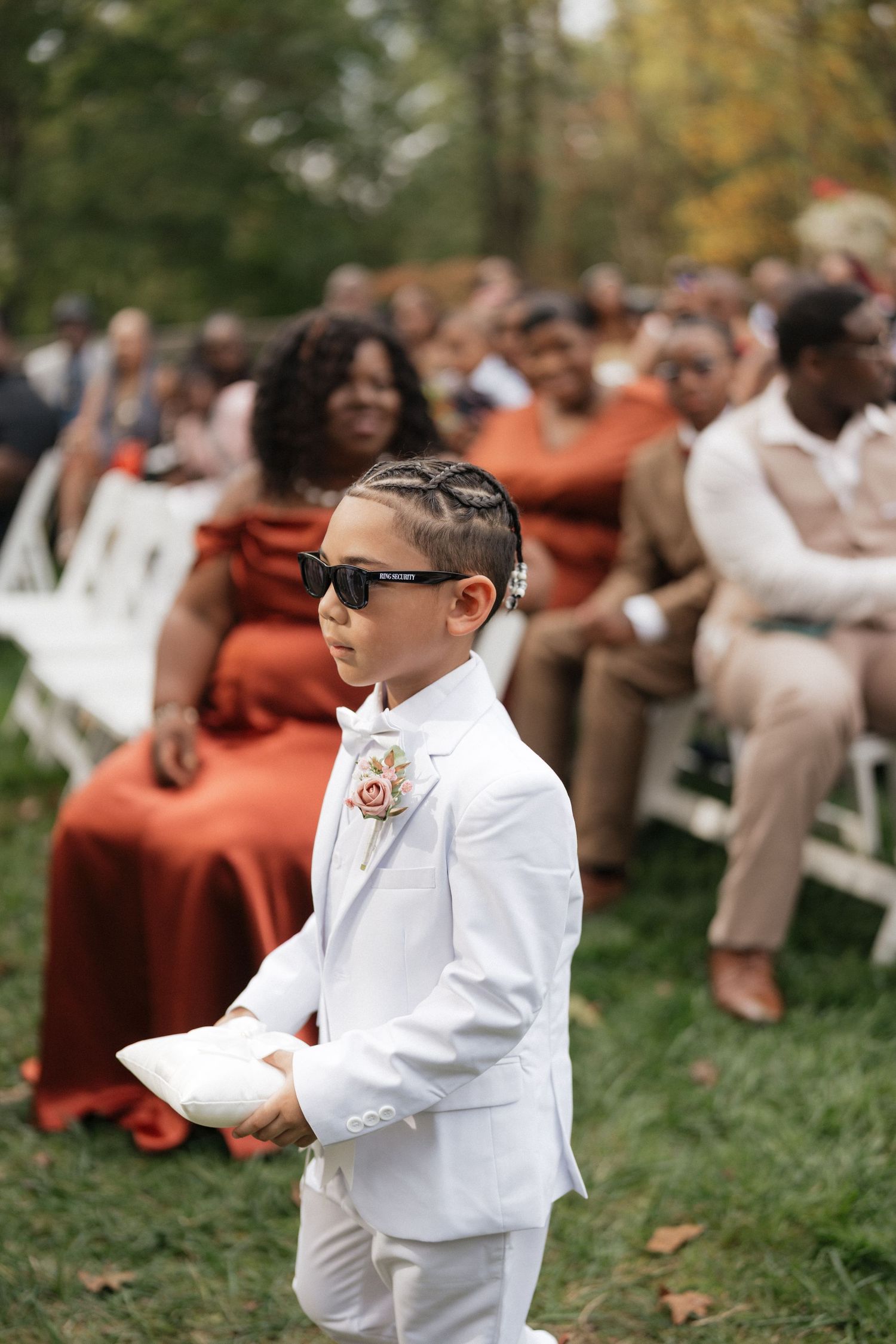 Young ring bearer in white suit dances enthusiastically at an outdoor wedding ceremony with guests in burgundy dresses.
