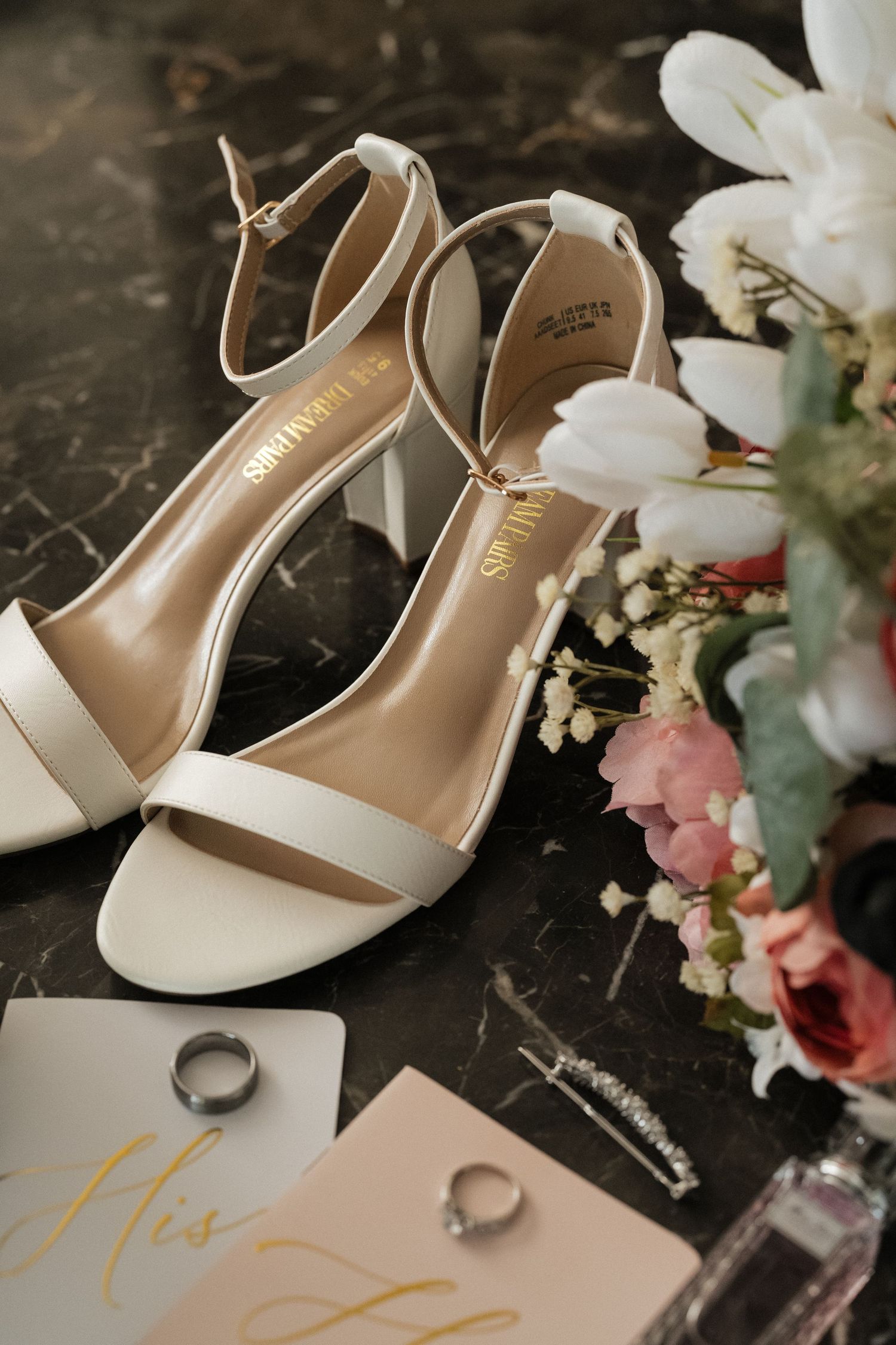 White bridal heels, wedding rings, and floral decorations arranged on a dark marble surface.