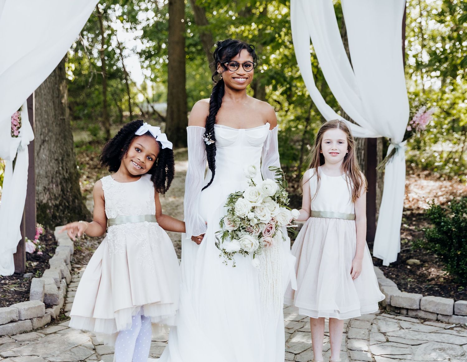 Wedding party members in white dresses pose together on stone pathway outdoors.