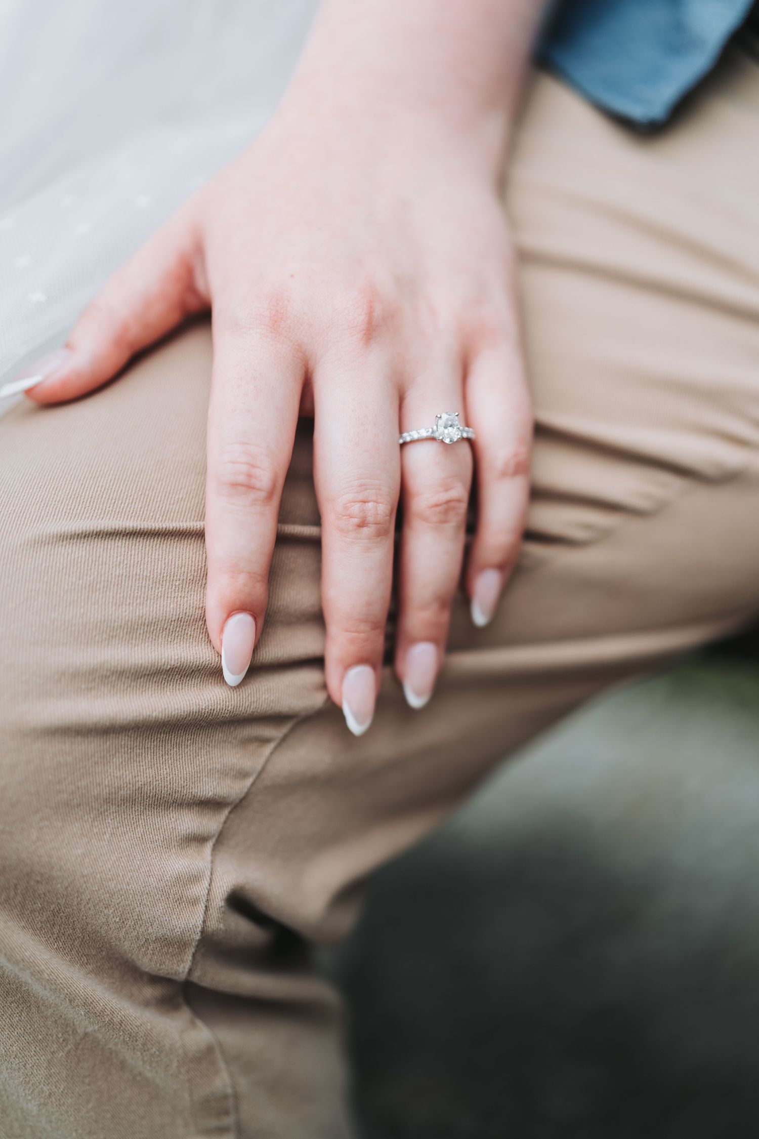 Close-up of a manicured hand with engagement ring resting on khaki fabric.