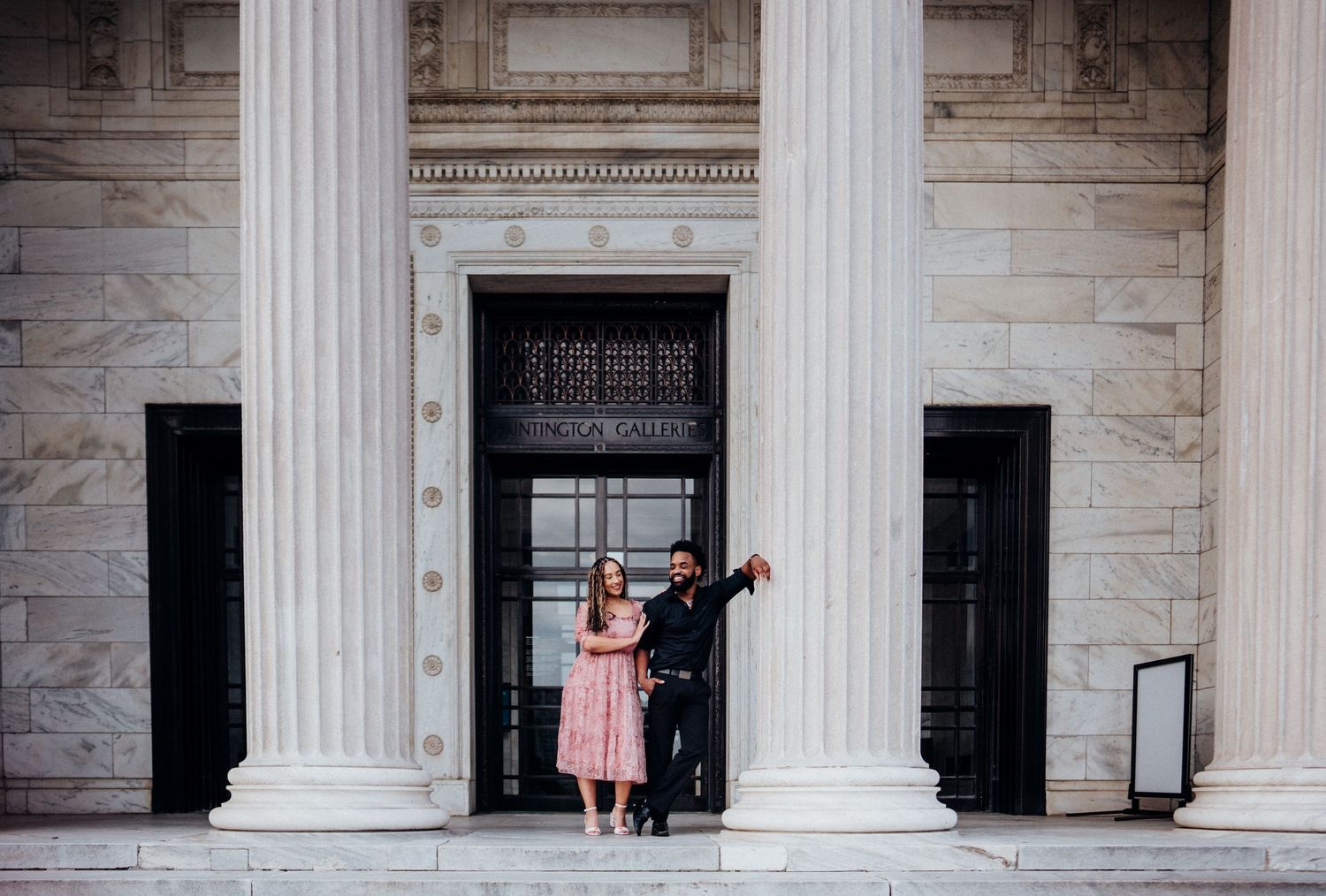 A romantic couple poses between grand white columns at an elegant building entrance.