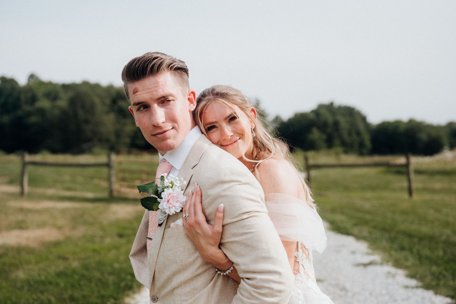 A couple in wedding attire poses together on a country path with green fields in the background.