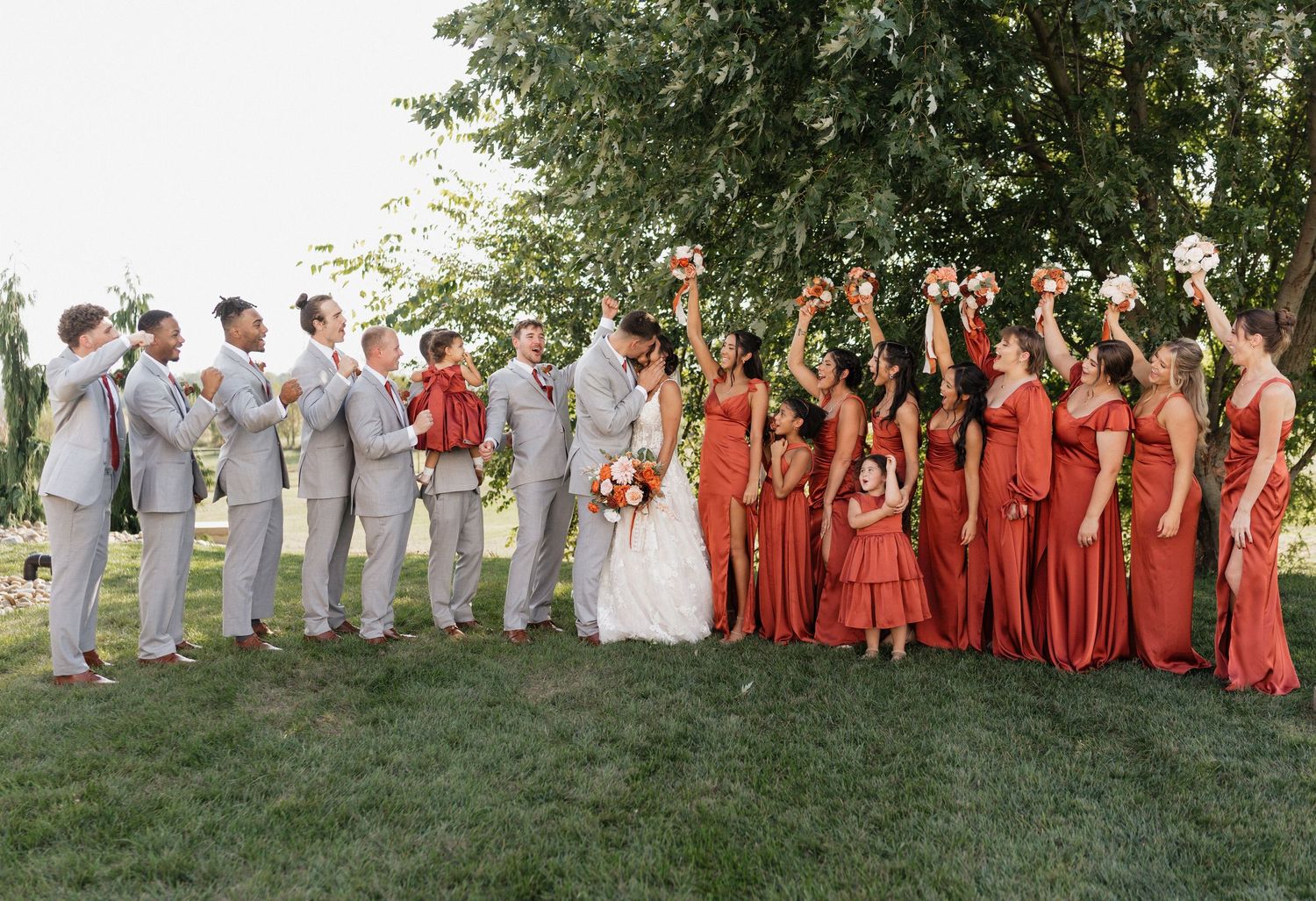 Large wedding party in grey suits and rust dresses posed for group photo on grassy hill.