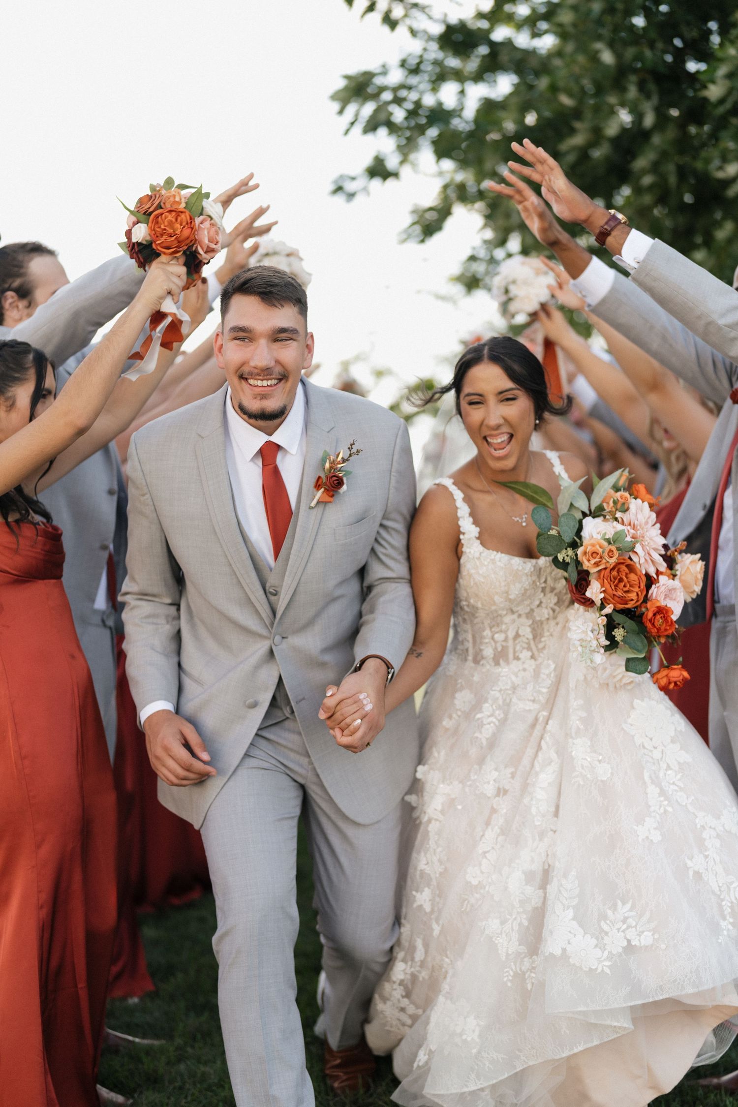 Joyful wedding party celebrating with arms raised outdoors in coral and grey attire.