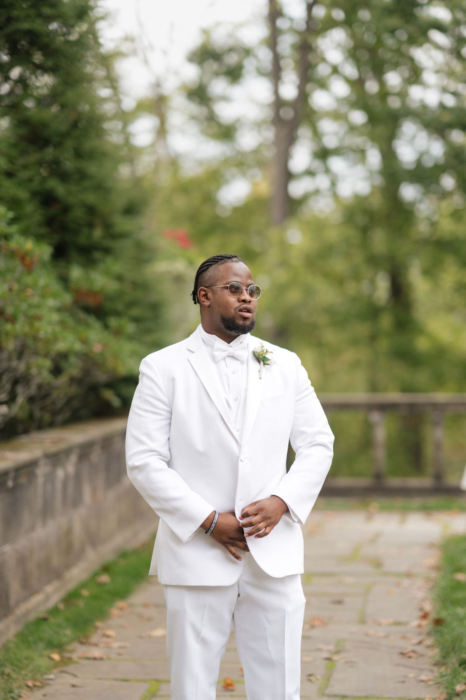 A person in an all-white formal suit poses outdoors in a garden setting.