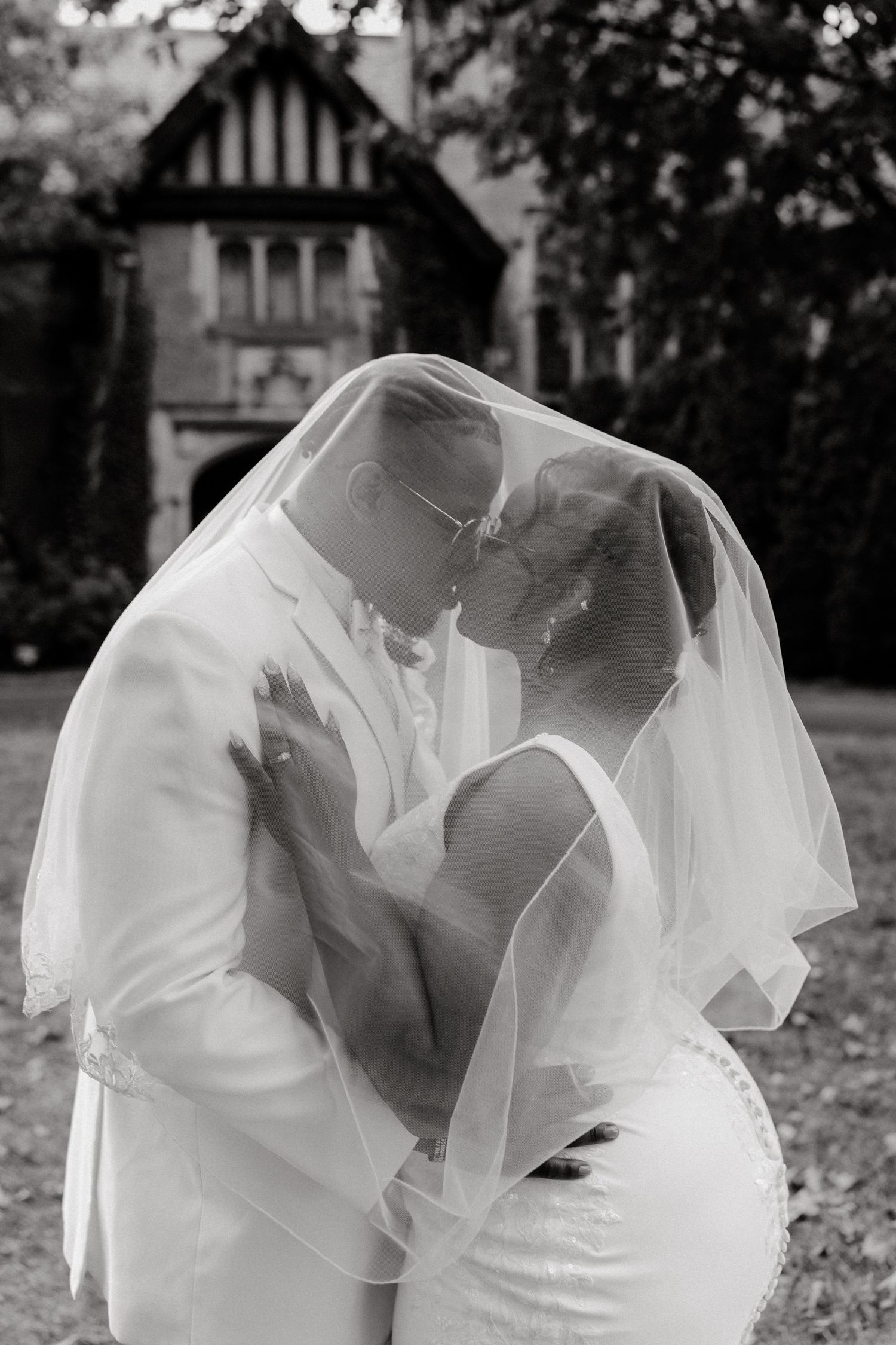 A tender black and white wedding portrait captured through a flowing veil outdoors.