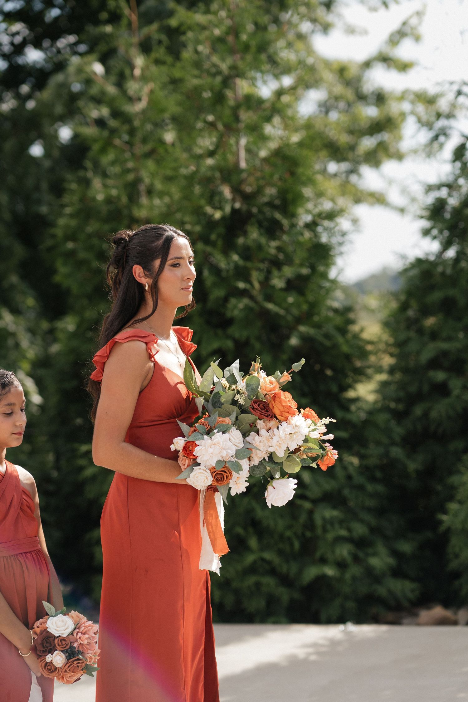 A bridesmaid in a coral dress holds a beautiful bouquet of white and orange flowers outdoors.