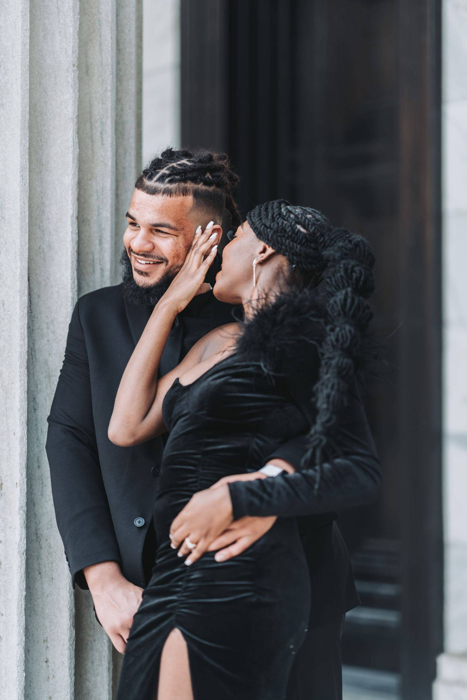 A couple shares a playful moment against a white pillared wall, dressed elegantly in black formal attire.