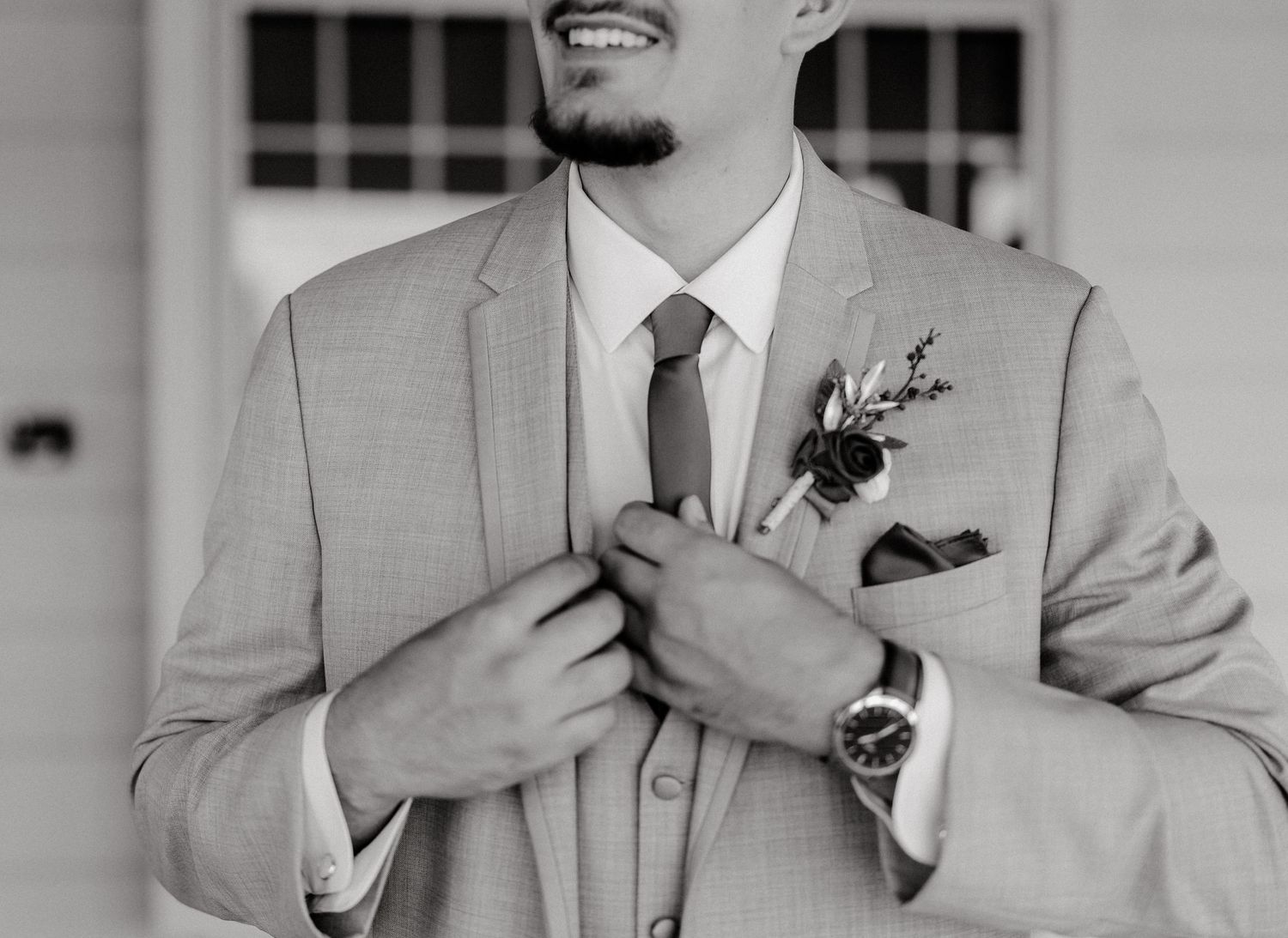 Groomsmen in light gray suits adjust their ties while smiling during wedding preparations.