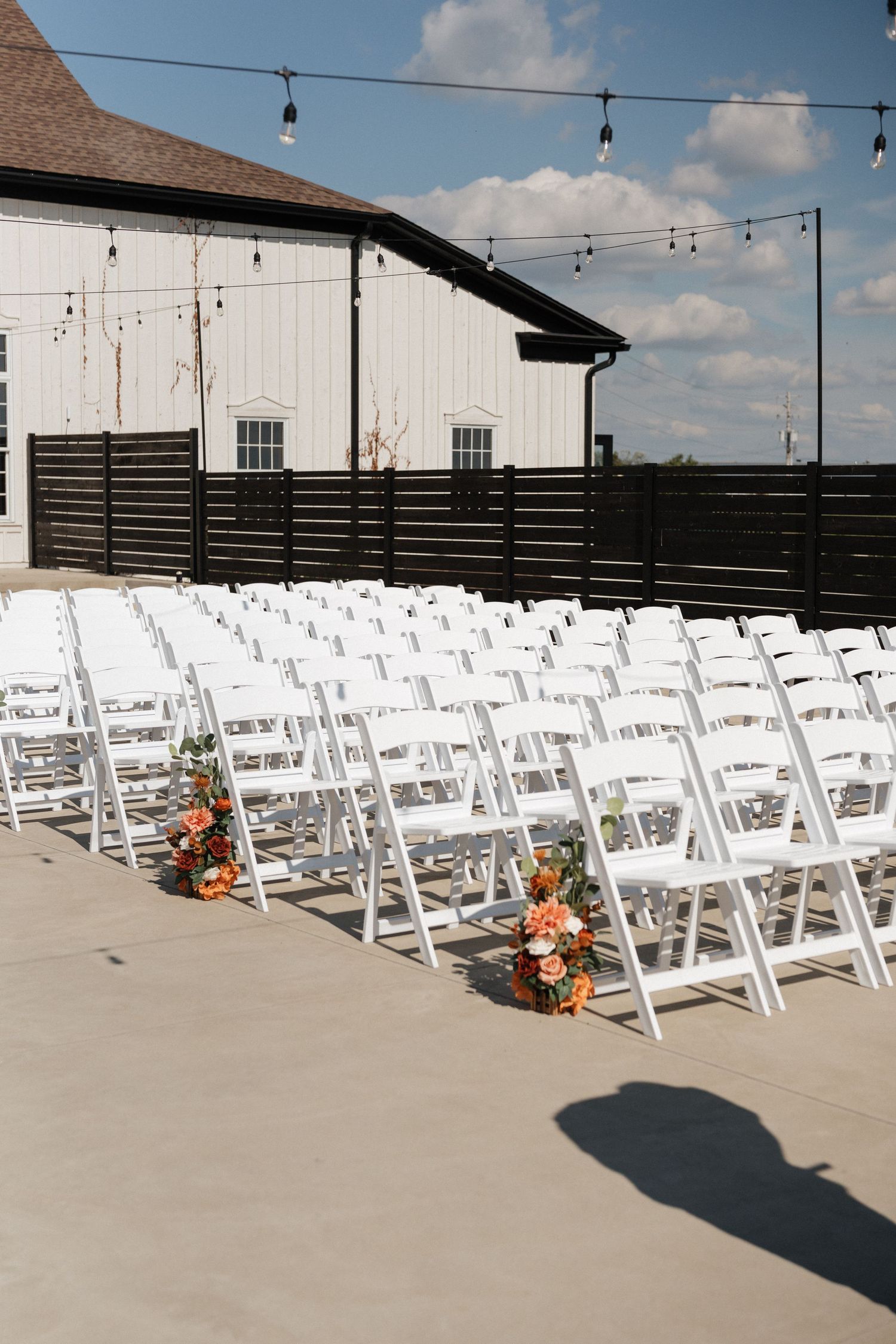 Outdoor wedding ceremony setup with white folding chairs and string lights against white barn backdrop.