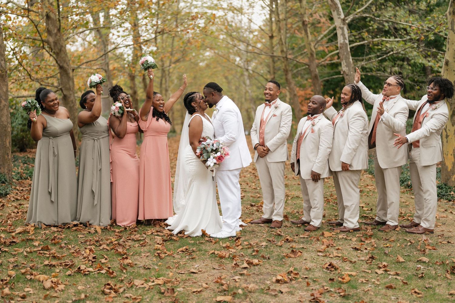 Wedding party poses in white and pink attire for group photo on autumn lawn surrounded by trees.