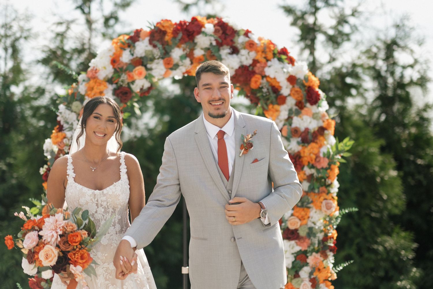 Joyful wedding couple celebrating under a floral arch with orange and white flowers.