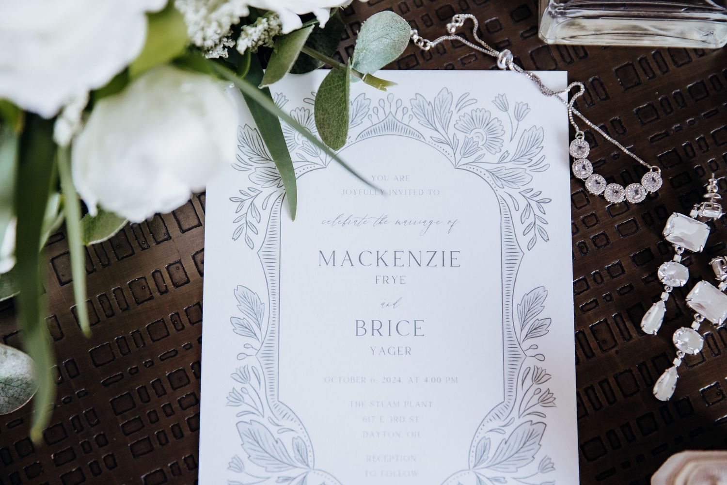Elegant white wedding invitation with floral border design displayed next to white roses and crystals.