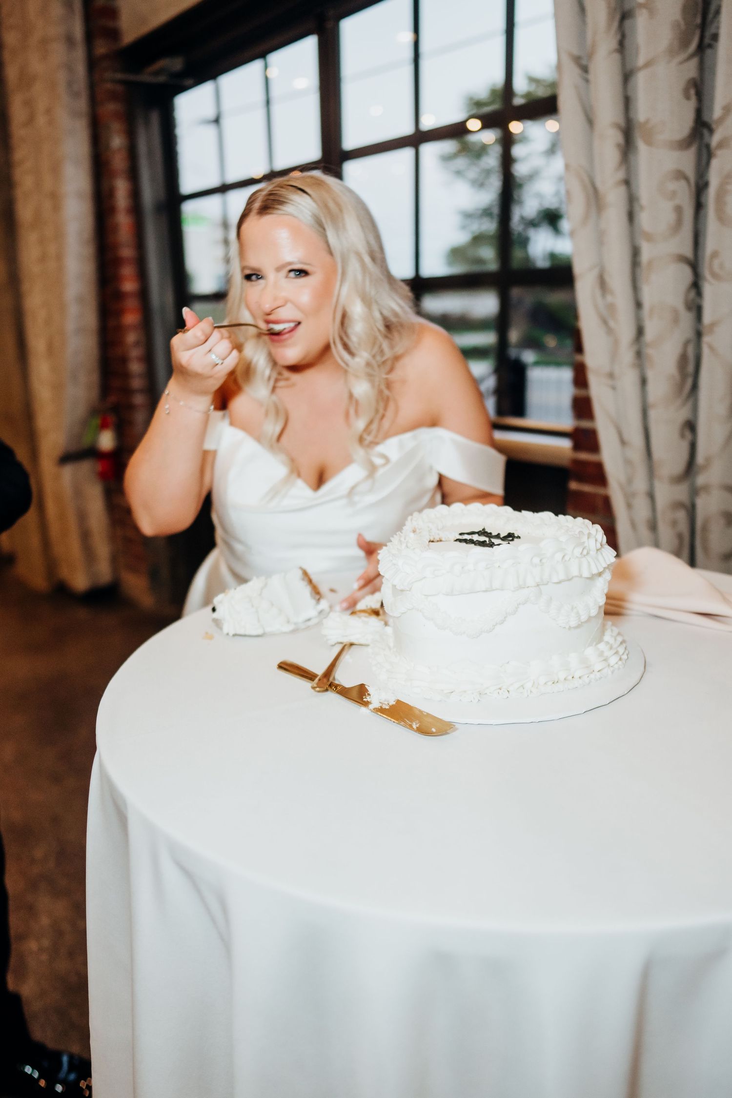 Bride in white dress enjoys wedding cake at reception table with elegant place settings.