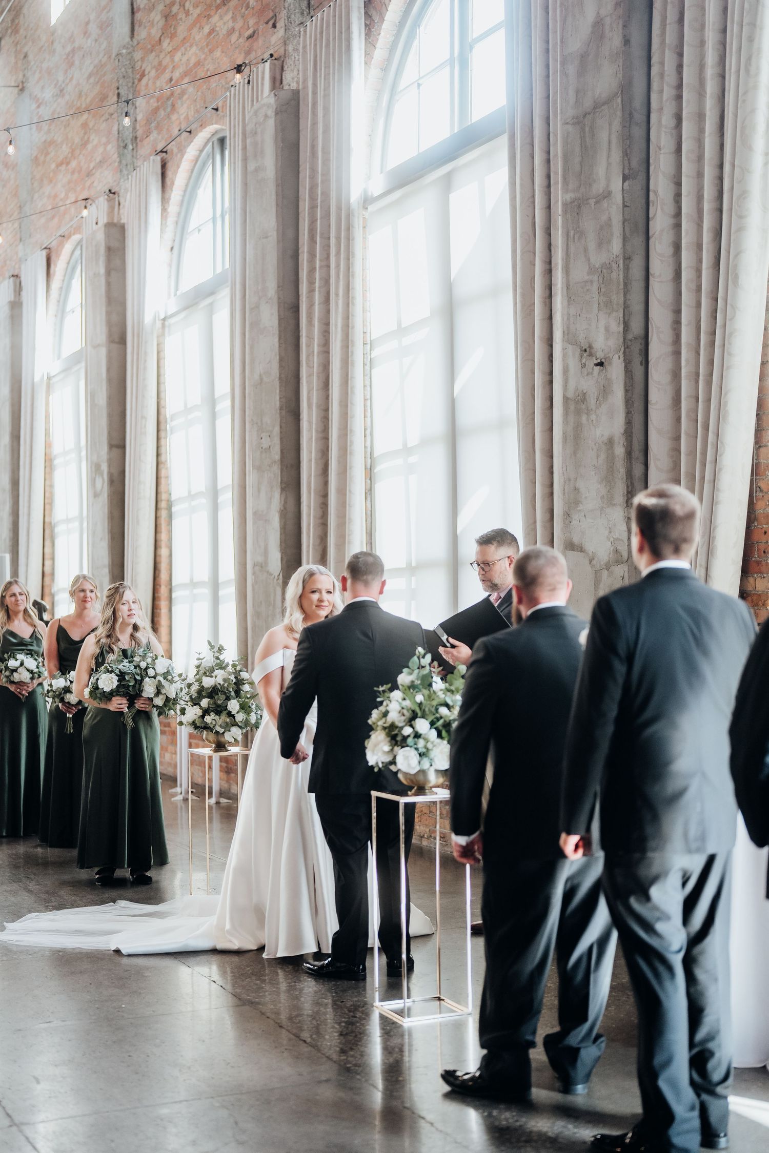 Wedding party lines up in an elegant indoor venue with tall windows and white floral arrangements.