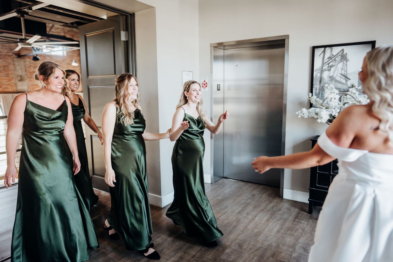 Wedding party members in emerald green dresses and white gown gather in an industrial-style hallway.