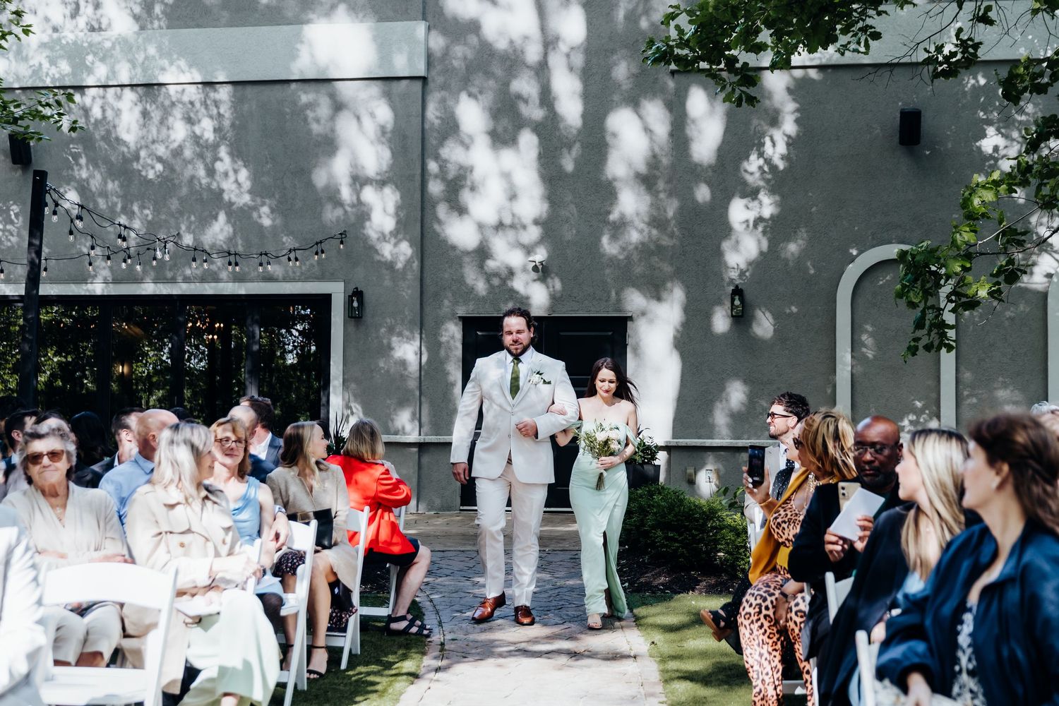 Wedding reception guests mingling outdoors under trees at venue.