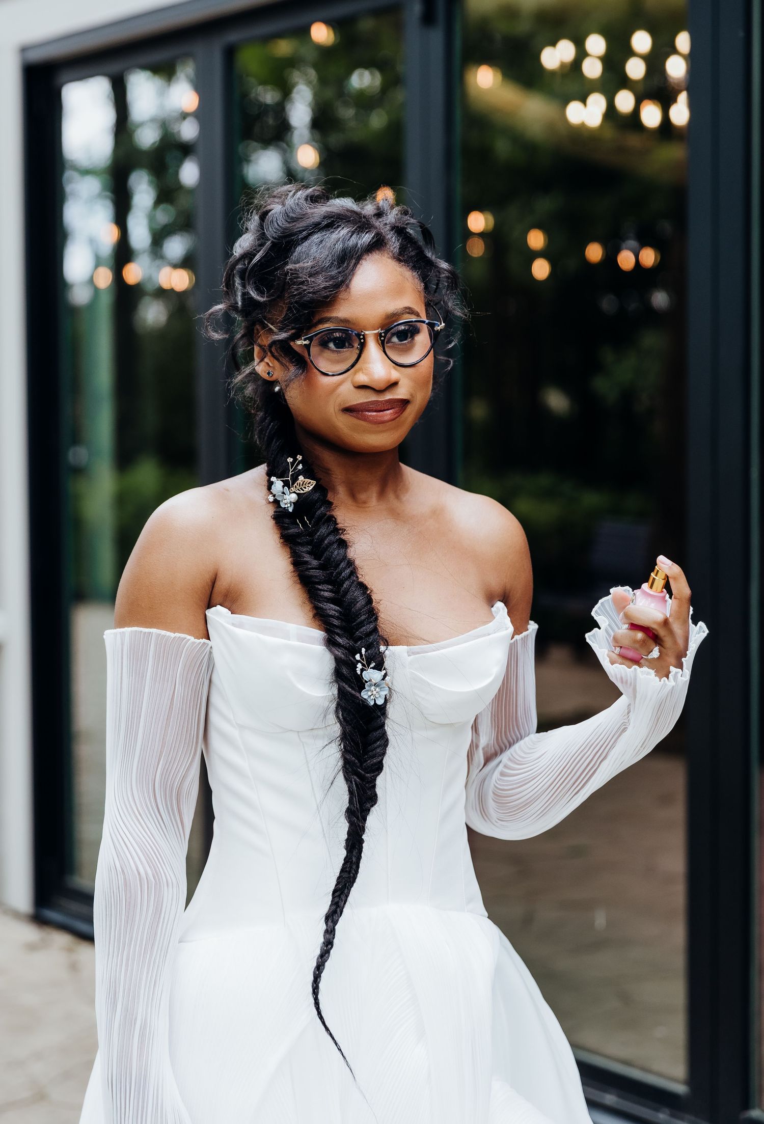A bride in an off-shoulder wedding dress with glasses and long braided hair poses against a modern building.