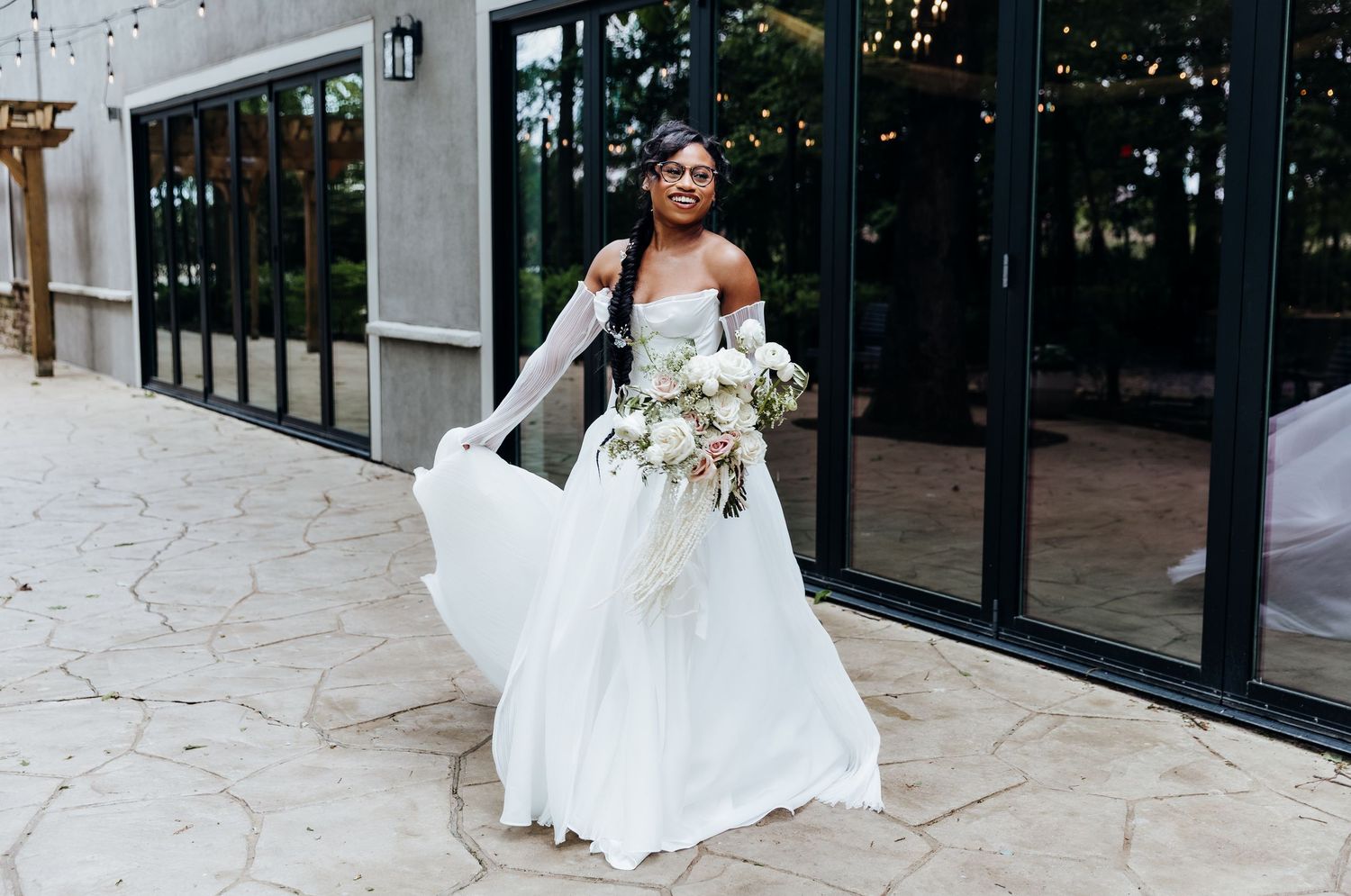 Bride in flowing white wedding gown posing against modern glass building exterior.