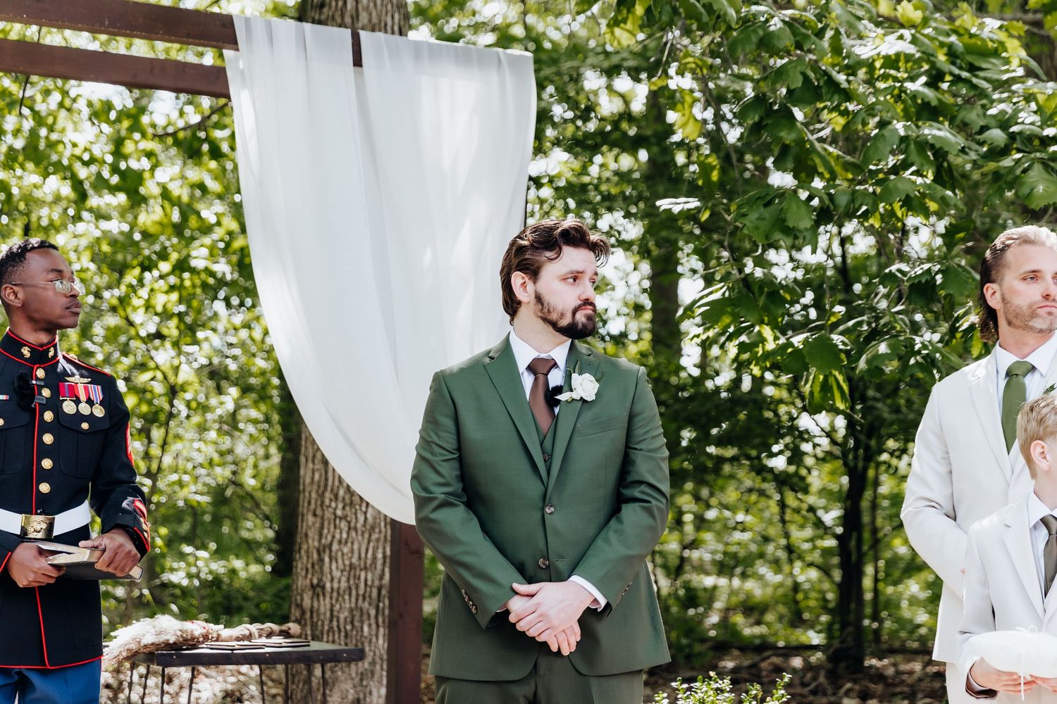 Wedding party members gathered outdoors with white ceremony chairs and greenery in background.