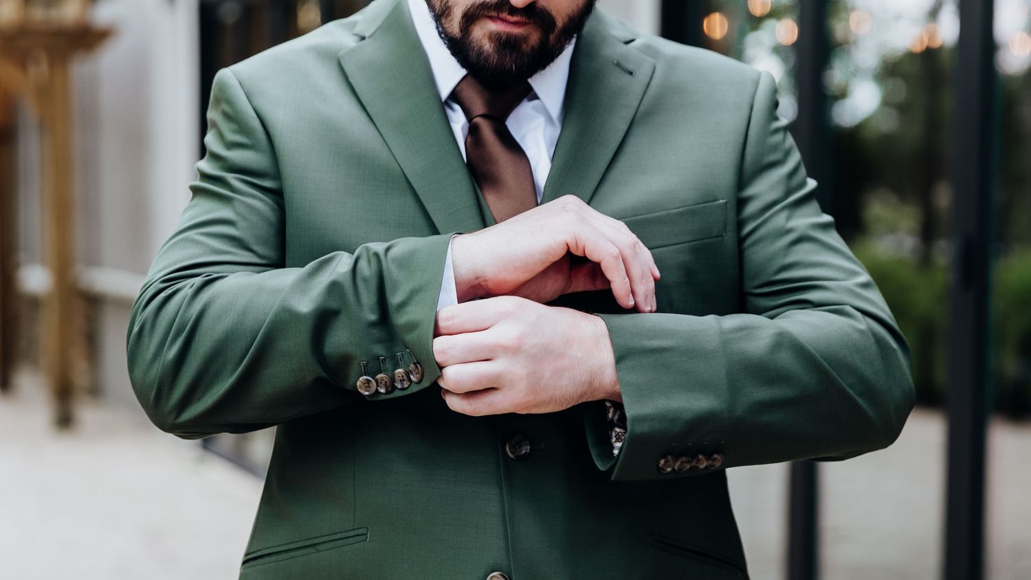A person in a green suit jacket checks their watch while standing in a well-lit hallway.